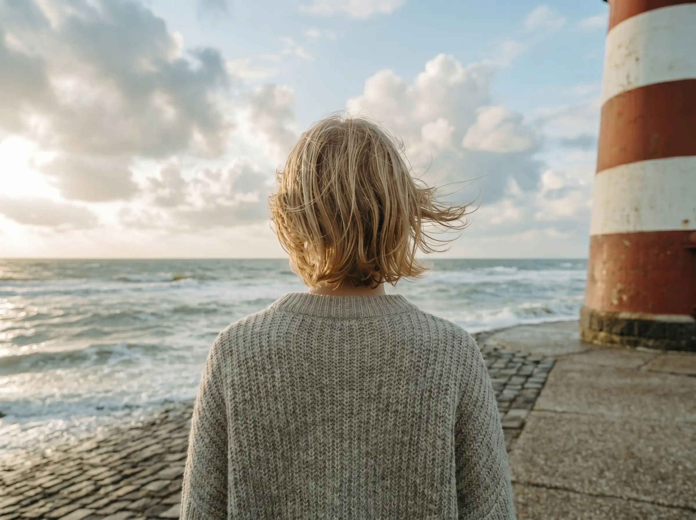 Person mit kurzen blonden Haaren und grauem Pullover blickt aufs Meer neben einem Leuchtturm.