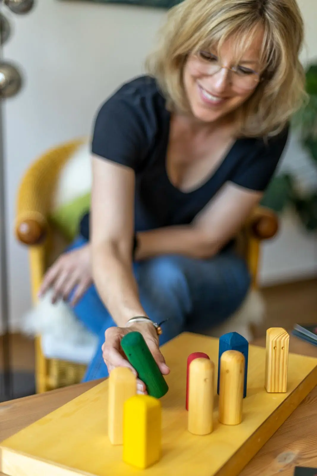 Frau mit Brille lächelt und bewegt grüne Holzfigur auf Spielbrett mit bunten Holzklötzen.