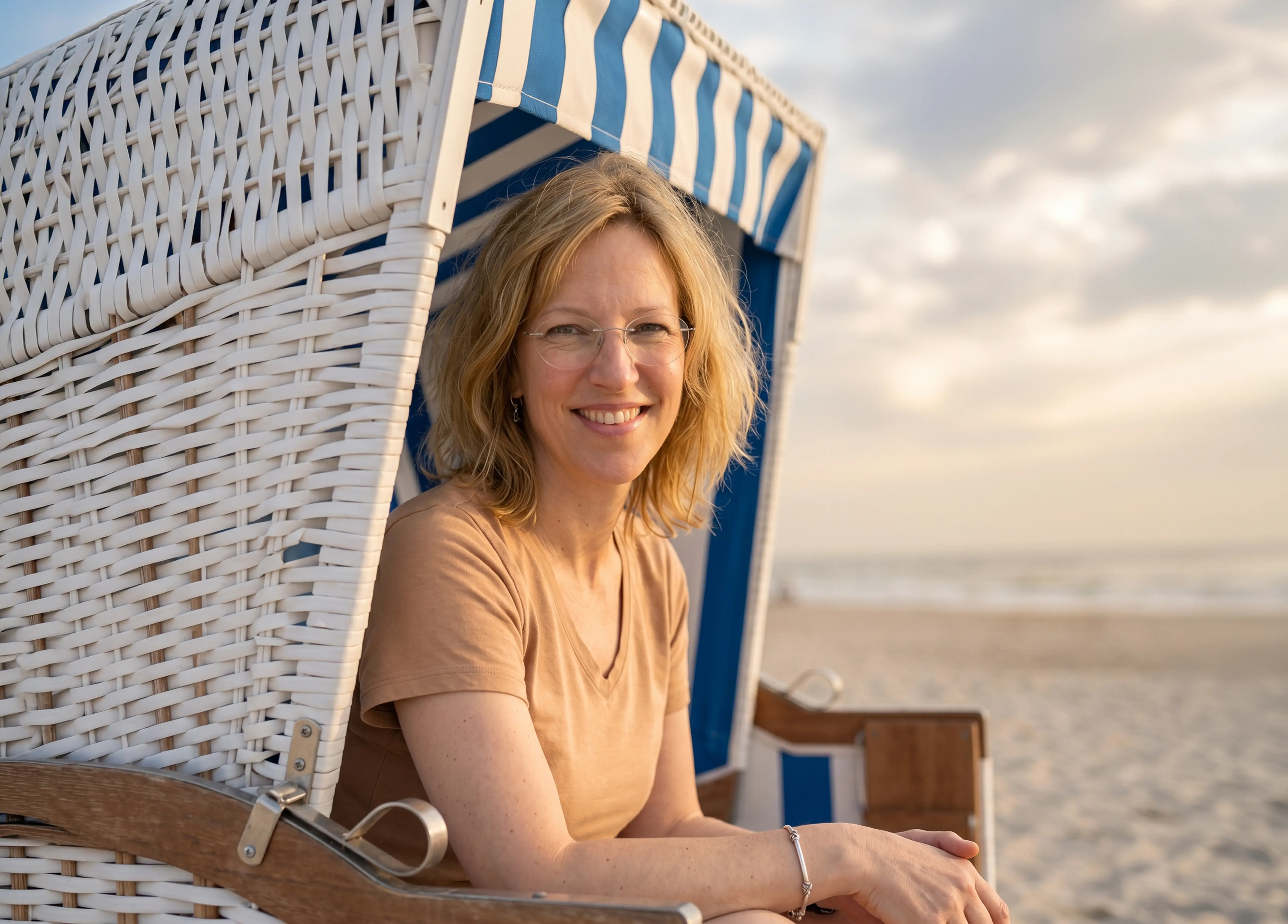 Lächelnde Frau mit Brille sitzt in einem weißen Strandkorb mit blauen Streifen am Strand.